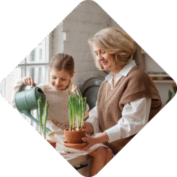 An photo of two people. On the left is a young girl holding a watering pitcher. On the right is an older woman holding a plant