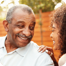 An photo of two people. On the left is an older man who is similing. On the right is an older woman looking at the man